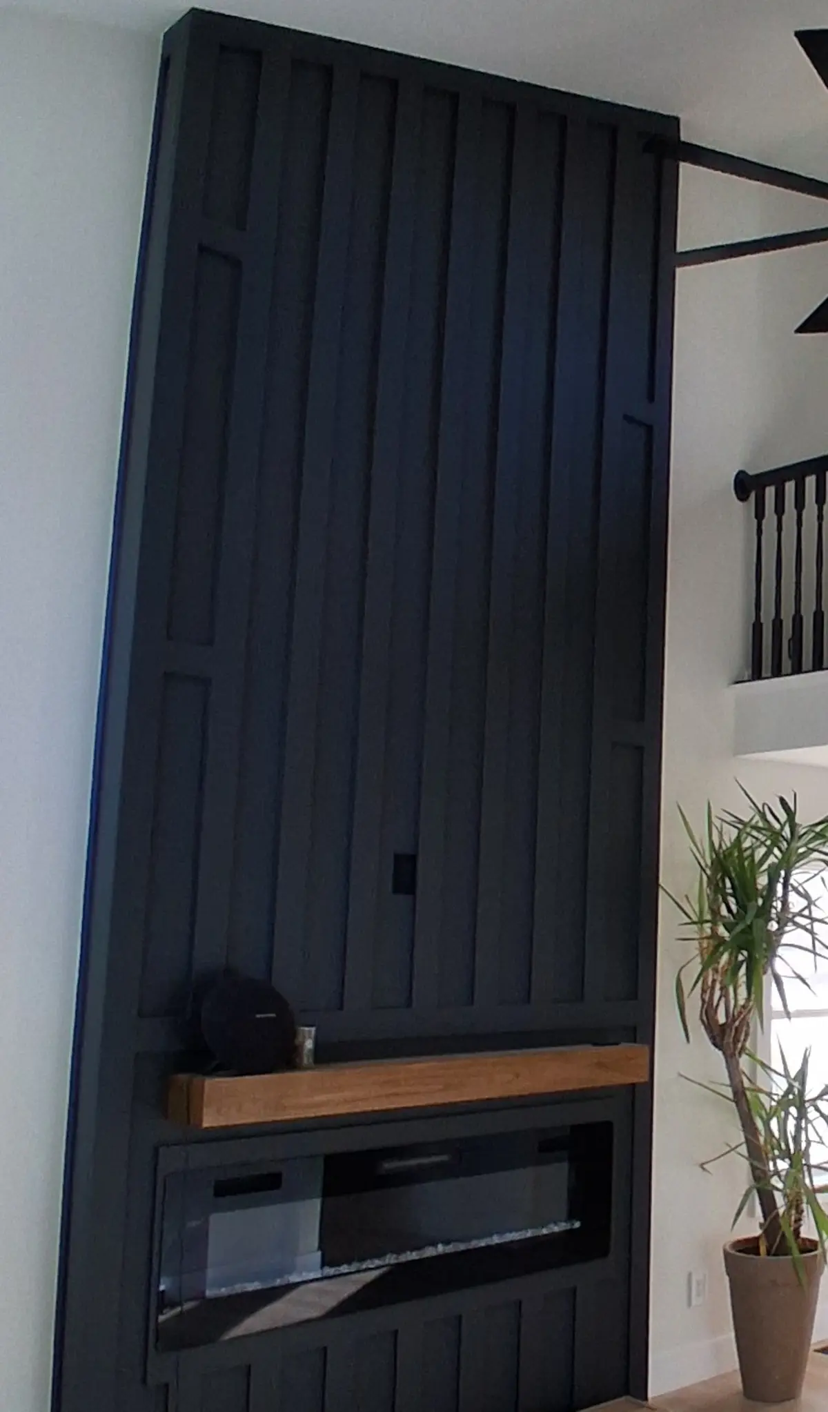 A two-storey black board-and-batten fireplace wall with a white oak mantel over a linear electric fireplace, a black loft rail beside it